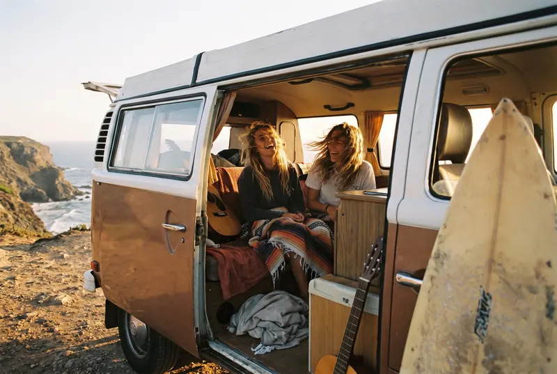 Freundinnen sitzen in einem offenen Campingbus an der Küste, daneben ein Surfbrett